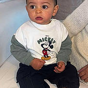 Manny a rejoint le concours — aidez-le/la à gagner de superbes lots ! toddler, child, mickey_mouse, sweatshirt, socks, black_pants, couch, white_couch, indoors, person, curly_hair, bracelet, knitted_sweater, adult_hand, seated, expression, large_eyes, casual_clothing, home, portrait