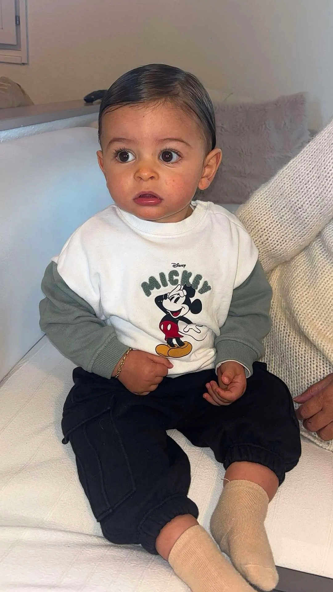 Manny a rejoint le concours — aidez-le/la à gagner de superbes lots ! toddler, child, mickey_mouse, sweatshirt, socks, black_pants, couch, white_couch, indoors, person, curly_hair, bracelet, knitted_sweater, adult_hand, seated, expression, large_eyes, casual_clothing, home, portrait