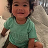 toddler, child, baby, smiling, curly_hair, portrait, sitting, bed, bedroom, blanket, clothing, green_outfit, indoors, window, blinds, happy, face, person, home, photograph