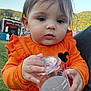 baby, background, child, closeup, clothing, curious, daylight, earrings, face, fair, grass, hand, holding, nature, orange_sweater, outdoor, person, plastic_cup, portrait, toddler