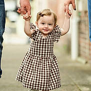 Ellie is registered to the contest to win money with this photo: toddler, child, smiling, walking, holding_hands, dress, checkered_pattern, white_shoes, outdoor, sidewalk, adult_hands, happy, cute, young_child, blonde_hair, hair_bow, person, family, footwear, support