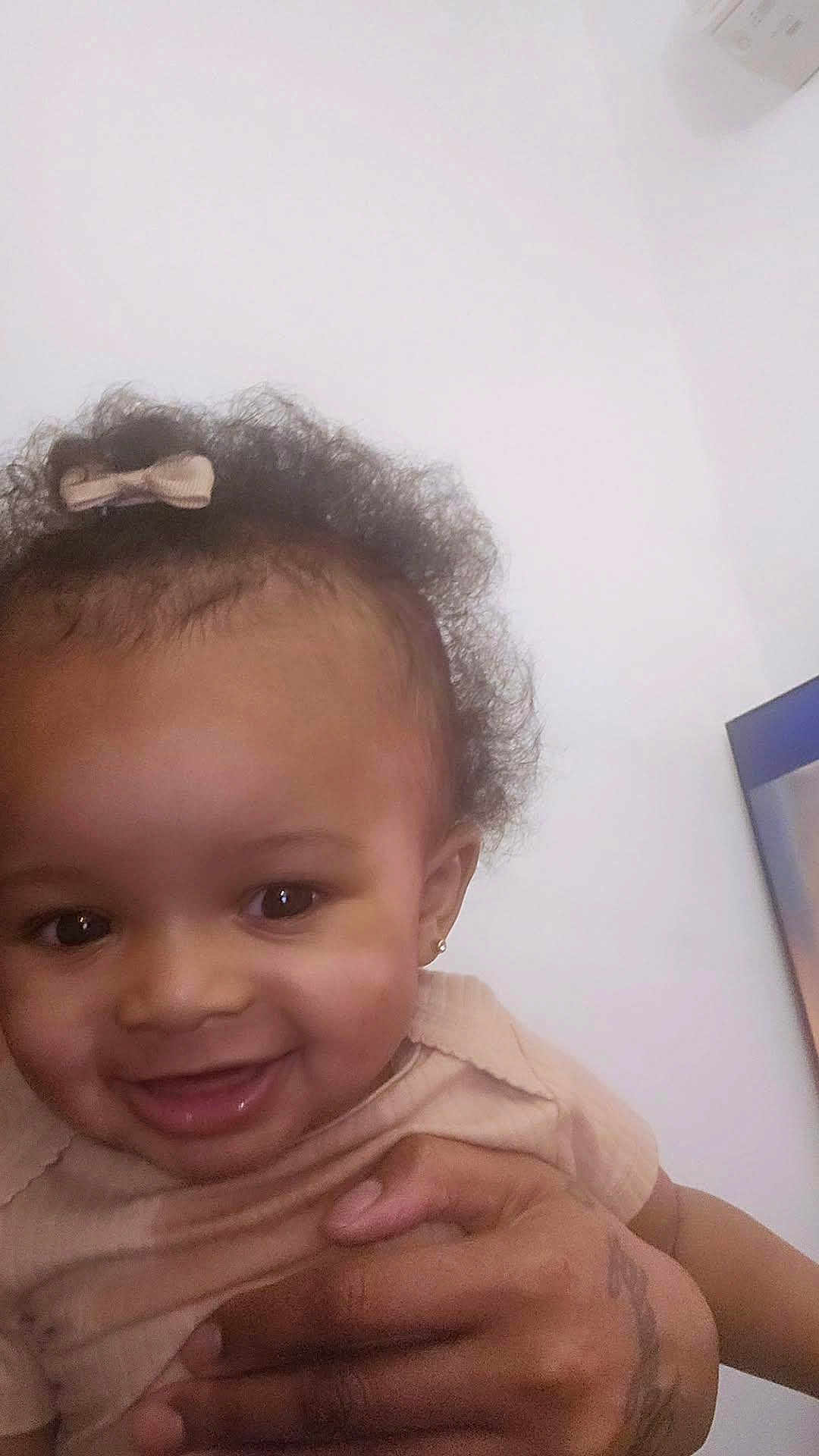 Lillie is registered to the contest to win money with this photo: baby, child, infant, smiling, portrait, closeup, curly_hair, hair_bow, earring, eyes, teeth, hand, holding, skin, indoor, white_wall, air_conditioner, cute, happy, person