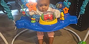 Lillie is registered to the contest to win money with this photo: baby, child, infant, face, eyes, hair, jumper, activity_center, toy, star_toy, fish_toy, frog_toy, stroller, couch, living_room, wooden_floor, feet, legs, seat, indoor