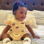 Amara is registered to the contest to win money with this photo: toddler, child, smile, curly_hair, knit_outfit, cherry_pattern, socks, bed, pillow, blanket, cozy, cute, happy, indoor, portrait, person, hair_tie, earrings, skin, seated