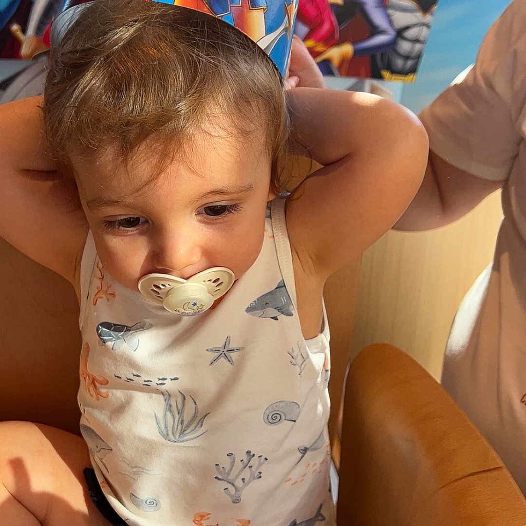 Hélio a rejoint le concours — aidez-le/la à gagner de superbes lots ! baby_outfit, celebration, child, costume, cute, decorations, face, high_chair, indoor, pacifier, paper_crown, party, person, seat_belt, smiling, superhero, toddler, wonder_woman, wooden_chair, young_child