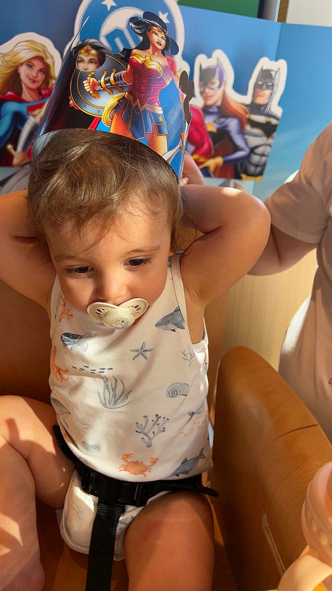 Hélio a rejoint le concours — aidez-le/la à gagner de superbes lots ! toddler, child, pacifier, high_chair, superhero, wonder_woman, paper_crown, costume, indoor, celebration, decorations, cute, smiling, young_child, seat_belt, wooden_chair, baby_outfit, face, person, party
