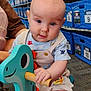 baby, child, toy, dinosaur_toy, rocking_toy, blue, storage_bins, indoor, carpet, person, cute, overalls, wooden_handle, play, infant, sitting, curious, expression, floor, store