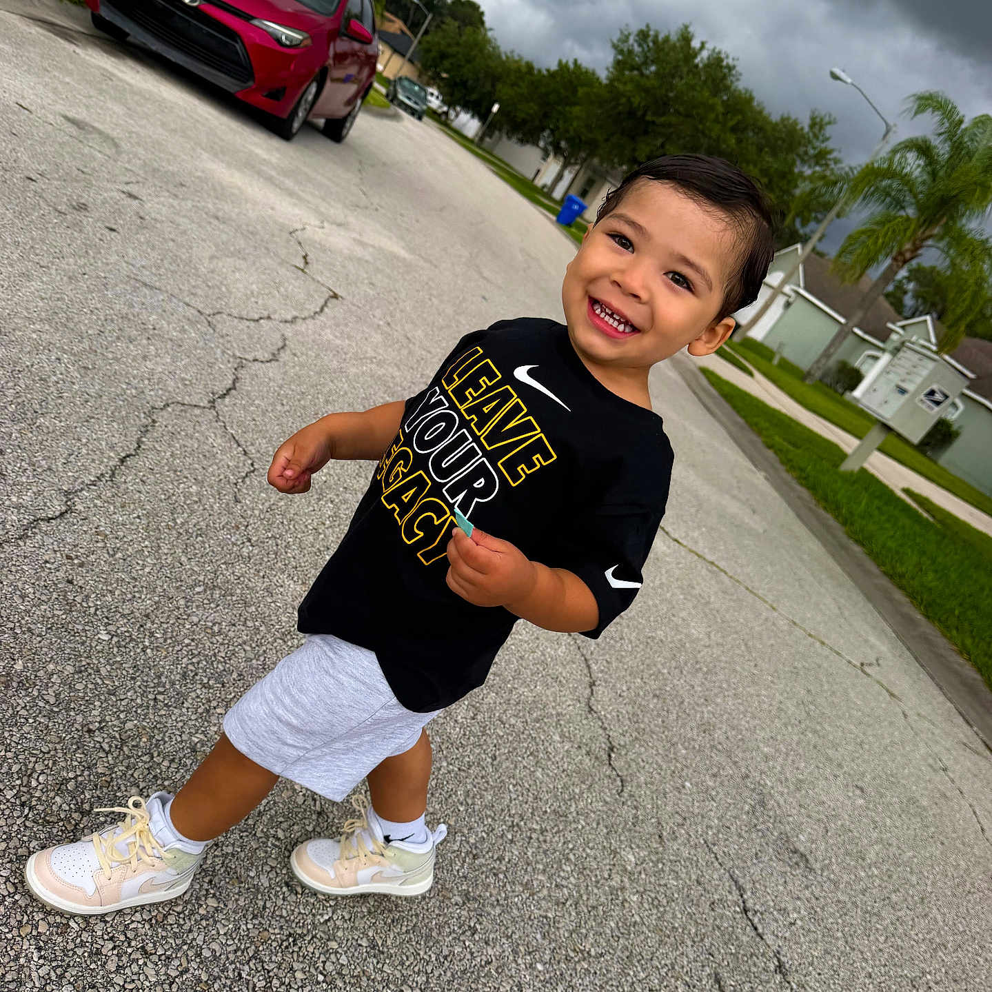 Liam Infante joined the competition — help win amazing prizes! toddler, child, smiling, street, sidewalk, car, red_car, tennis_shoes, shorts, black_shirt, text_on_shirt, cloudy_sky, trees, grass, residential_area, palm_tree, asphalt, walking, happy, outdoor