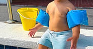 Liam Infante is registered to the contest to win money with this photo: toddler, child, swimming_pool, water, floaties, yellow_bucket, curly_hair, shorts, sunlight, outdoor, sitting, feet_in_water, play, summer, daytime, person, skin, happy, relaxation, fun