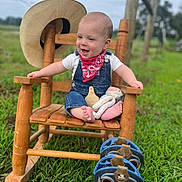 Lane joined the competition — help win amazing prizes! baby, child, rocking_chair, outdoor, grass, hat, denim_overalls, bandana, toy, footwear, cowboy_boots, wood, smiling, happy, person, nature, plush_toy, feet, countryside, portrait