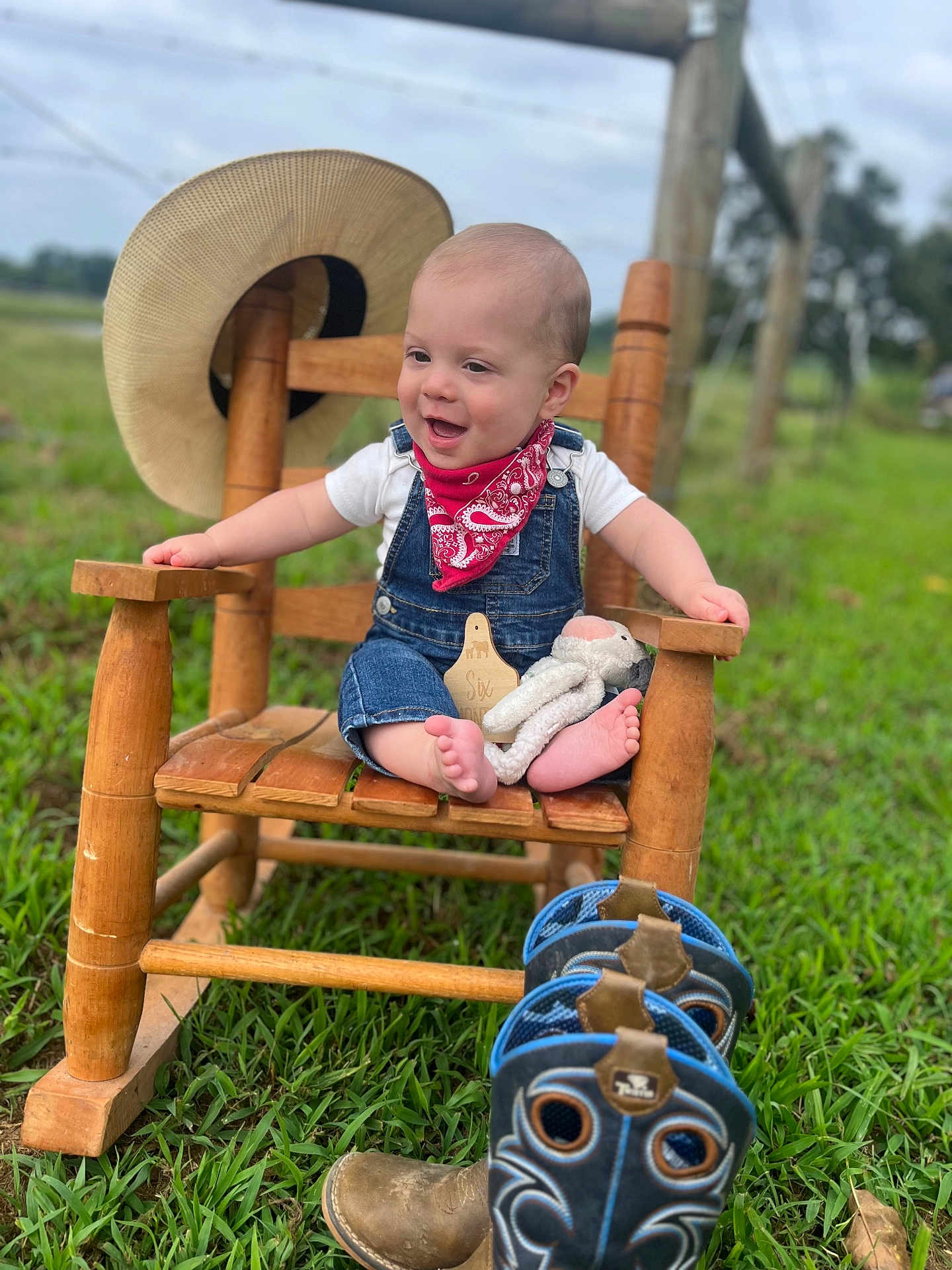 Lane joined the competition — help win amazing prizes! baby, child, rocking_chair, outdoor, grass, hat, denim_overalls, bandana, toy, footwear, cowboy_boots, wood, smiling, happy, person, nature, plush_toy, feet, countryside, portrait