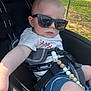 baby, child, sunglasses, stroller, outdoor, sunlight, clothing, shorts, tshirt, seatbelt, beads, hand, face, head, person, summer, shade, fence, grass, reflection