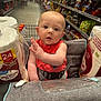 baby, child, shopping_cart, toilet_paper, supermarket, aisle, snacks, red_clothing, face, hand, grocery_store, retail, plastic_wrap, seatbelt, curious, indoor, person, shopping, product_packaging, store_shelves