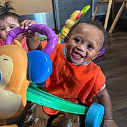 Nyzaun joined the competition — help win amazing prizes! baby, child, colorful, curious, door, face, floor, furniture, hand, happy, high_chair, indoor, orange_bib, pants, play, playful, smile, toddler, toy, wood_floor