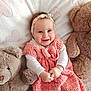 baby, smiling, pink_dress, headband, teddy_bear, plush_toy, blanket, heart_pattern, infant, cute, happy, soft_texture, child, portrait, indoors, cozy, white_long_sleeve, cuddle, toy, comfort