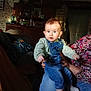 Olivia participe au concours pour gagner de l'argent avec cette photo : baby, child, denim_overalls, sweater, socks, adult_hand, living_room, stone_wall, wooden_cabinet, clock, cushion, sofa, person, indoor, curious, blue_eyes, casual_clothing, patterned_shirt, holding, portrait