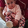 baby, child, infant, person, adult, holding, pink_clothing, floral_leggings, white_booties, fuzzy_rug, indoor, casual, seated, curious_expression, hand, legs, face, shorts, maroon_shirt, comfort