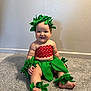 baby, child, smiling, costume, leaf_accessories, red_polka_dot, green_leaves, carpet, indoor, cute, sitting, barefoot, wall, adorable, headwear, wristbands, ankle_bands, plush, portrait, happy