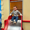 child, face, hair, handrail, happy, indoor_playground, infant, jeans, metal_slide, pipe, play, play_structure, portrait, red, skin, slide, smile, socks, sweater, toddler