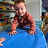 adult_partial, baby, blue_mat, child, clothing, crawling, face, foam_mat, hands, indoor, pacifier, person, play_area, playroom, portrait, shelf, smiling, toddler, toy_truck, toys