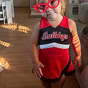 Arizona is registered to the contest to win money with this photo: barefoot, bulldogs, casual, cheerleading_outfit, child, clothing, couch, curious_expression, furniture, glasses, home, indoor, natural_light, person, portrait, red_glasses, smile, toddler, wooden_floor, young_child