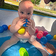 Atlas is registered to the contest to win money with this photo: baby, child, pool, inflatable_pool, plastic_balls, blue_ball, yellow_ball, pink_ball, water, diaper, hand, face, outdoor, grass, tree, building, sunlight, playing, cute, summer