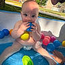 baby, blue_ball, building, child, cute, diaper, face, grass, hand, inflatable_pool, outdoor, pink_ball, plastic_balls, playing, pool, summer, sunlight, tree, water, yellow_ball