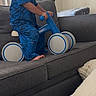 toddler, child, blue_pajamas, ride_on_toy, couch, gray_couch, cushion, indoor, living_room, smiling, playful, toy, barefoot, person, furniture, window_blinds, happy, young_child, casual, home
