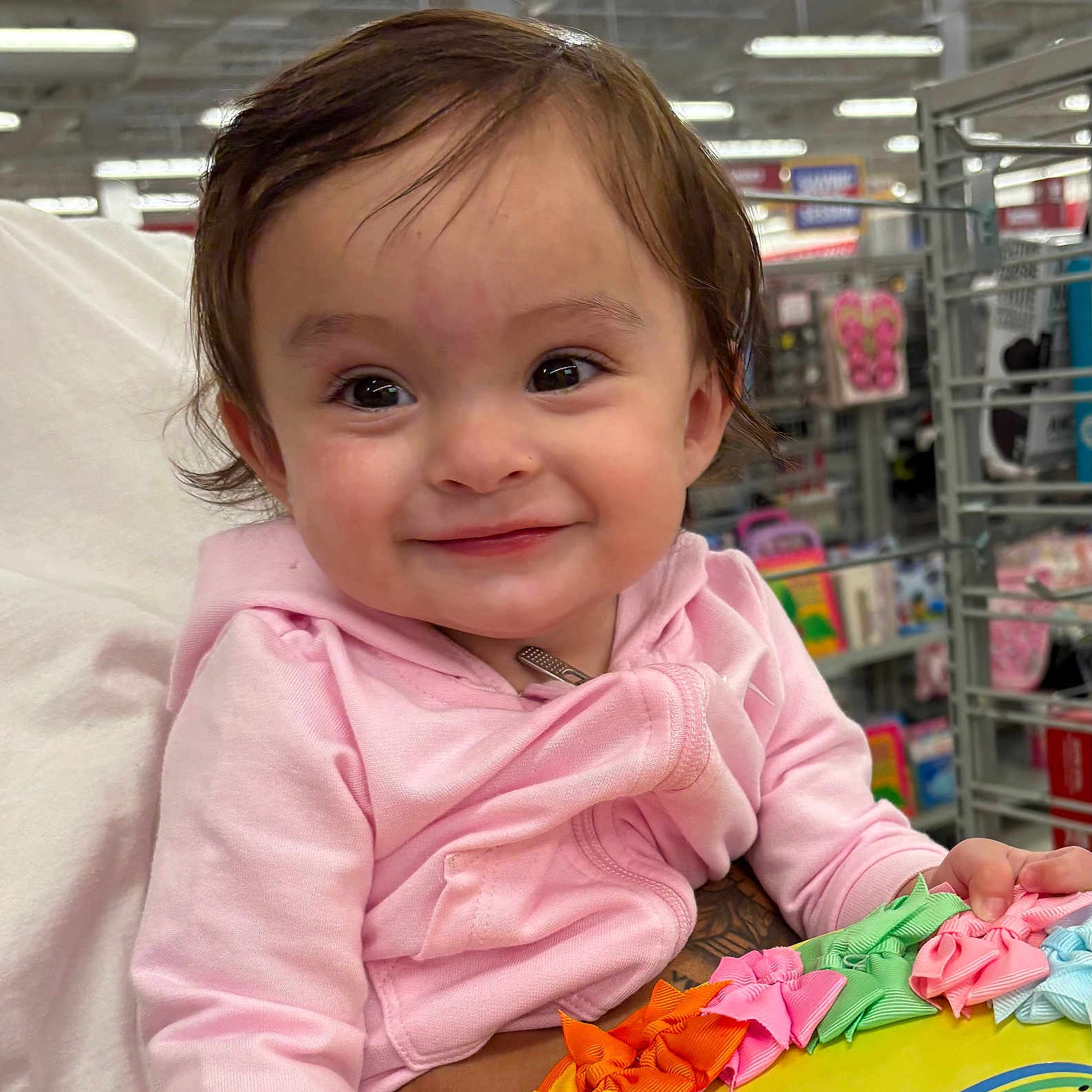 Galilea is registered to the contest to win money with this photo: aisle, baby, child, clothing, colorful_ribbons, cute, face, hand, happy, holding, indoor, person, pink_hoodie, retail, shelf, shopping, sleeve, smile, store, toy