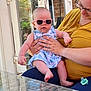 Annabelle is registered to the contest to win money with this photo: baby, sunglasses, adult, woman, holding, dress, patterned_clothing, glasses, smiling, outdoor, table, glass_table, reflection, hand, skin, person, seated, pacifier, window, door