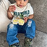 casual_clothing, child, couch, curly_hair, cute, food_container, green_shoes, indoor, jeans, living_room, looking_up, pillows, relaxed, ripped_jeans, shirt, sitting, snack, sneakers, toddler, wild_world_shirt