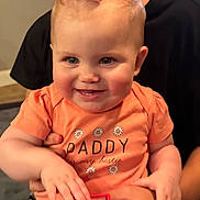 Kaylee is registered to the contest to win money with this photo: baby, bestie, child, closeup, clothing, cute, daddy, face, hands, happy, human, indoor, infant, orange_shirt, person_holding, pink_bow, portrait, smile, toy, young_child