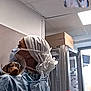 person, dog, medical_scrubs, protective_cone, veterinary, clinic, cage, face_mask, head_covering, video_call, indoor, table, spray_bottle, box, window, animal_care, looking_away, female, concerned, sitting
