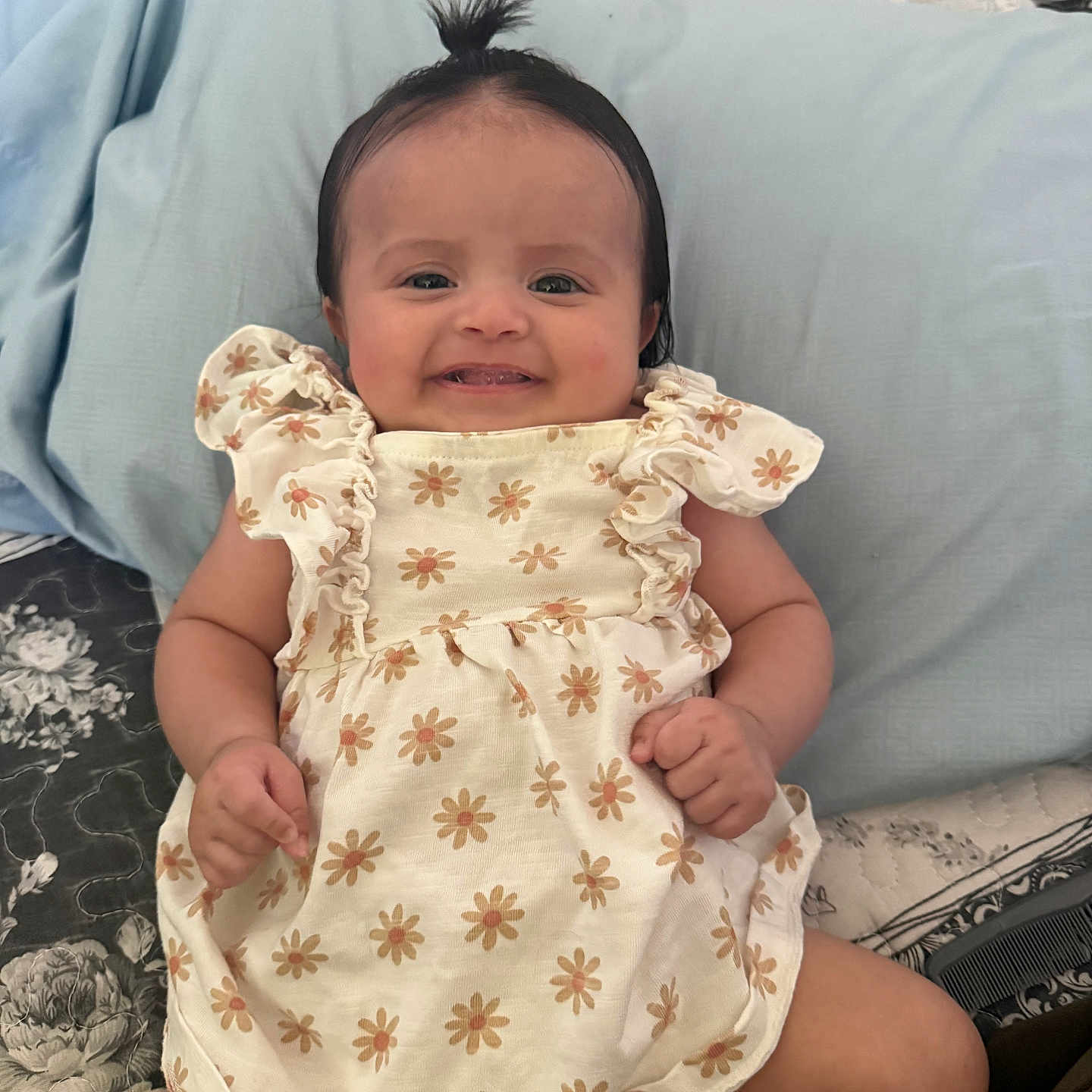 Jazlyn is registered to the contest to win money with this photo: baby, child, smiling, dress, flower_pattern, bed, pillow, cute, happy, indoors, person, infant, hair_tie, pattern, fabric, skin, face, small_hands, lying_down, comfort