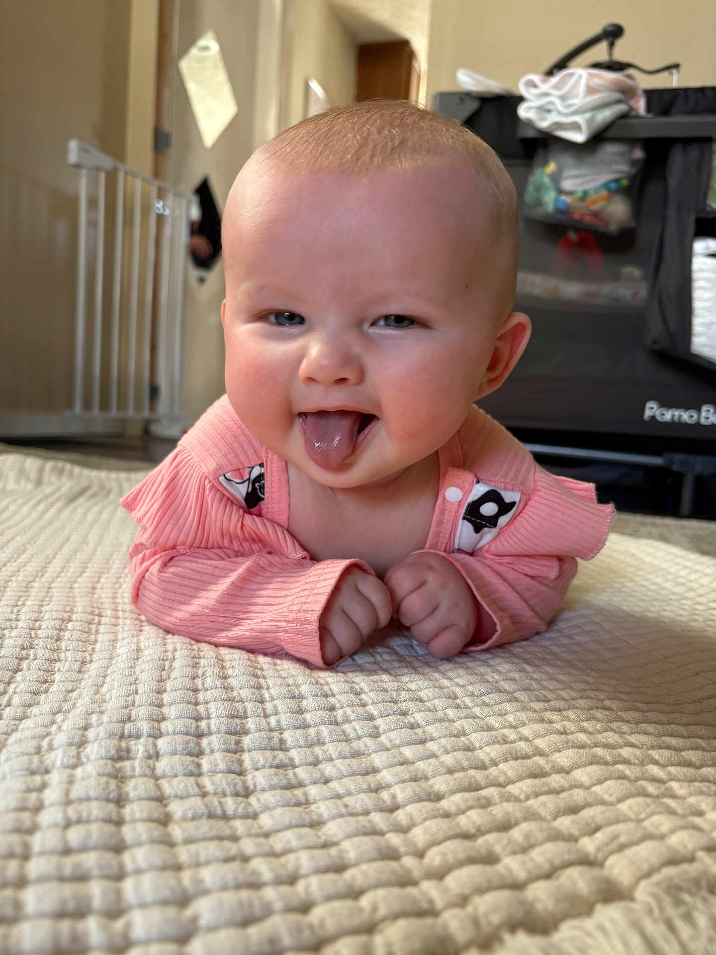 Emma is registered to the contest to win money with this photo: baby, tongue, pink_clothing, blanket, indoor, playpen, baby_gate, floor, person, child, cute, smiling, face, hands, head, expression, infant, home, cozy, texture