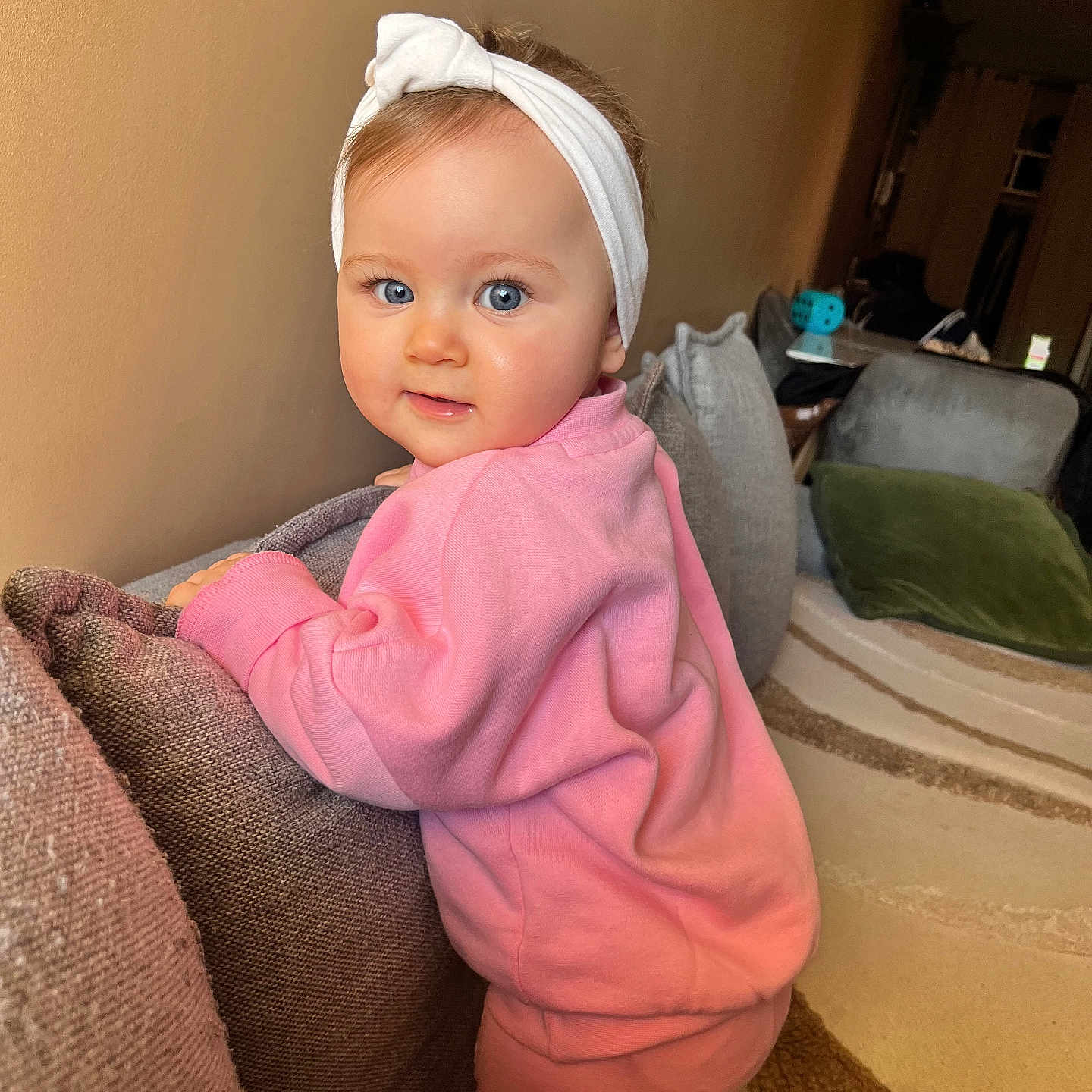 Jena participe au concours pour gagner de l'argent avec cette photo : baby, child, pink_clothing, headband, blue_eyes, couch, cushion, indoor, carpet, curious, portrait, person, toddler, warm_lighting, home, soft_fabric, face, smiling, standing, cozy