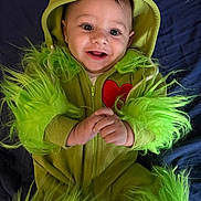 Marche is registered to the contest to win money with this photo: baby, child, costume, green, fuzzy, hood, smile, happy, face, hands, red_heart, cute, infant, lying_down, apparel, portrait, bright_eyes, fabric, playful, indoor