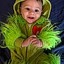 baby, child, costume, green, fuzzy, hood, smile, happy, face, hands, red_heart, cute, infant, lying_down, apparel, portrait, bright_eyes, fabric, playful, indoor