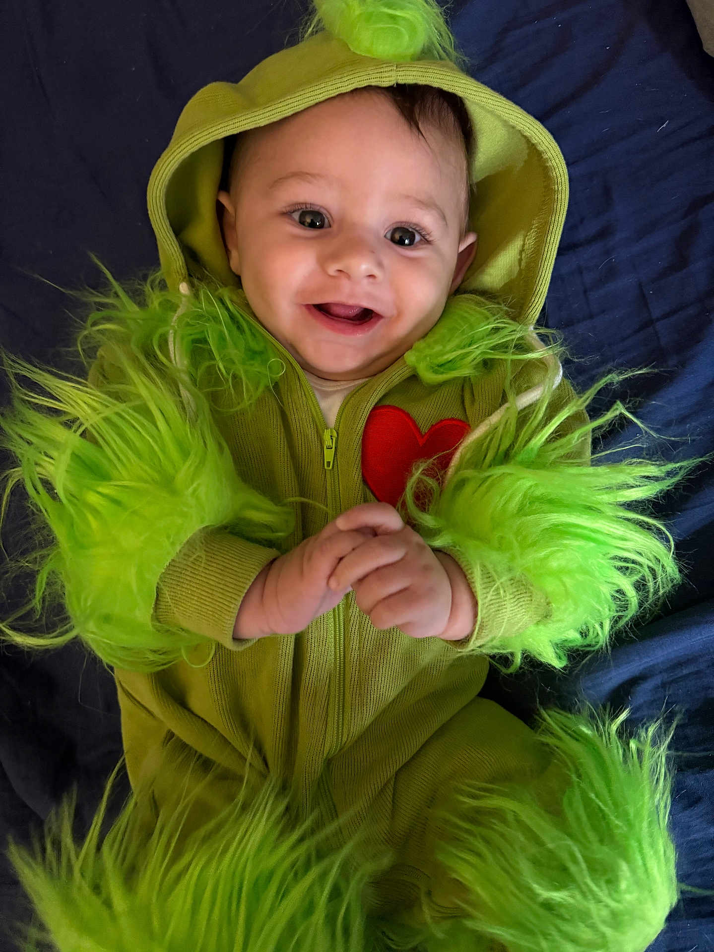Marche is registered to the contest to win money with this photo: baby, child, costume, green, fuzzy, hood, smile, happy, face, hands, red_heart, cute, infant, lying_down, apparel, portrait, bright_eyes, fabric, playful, indoor