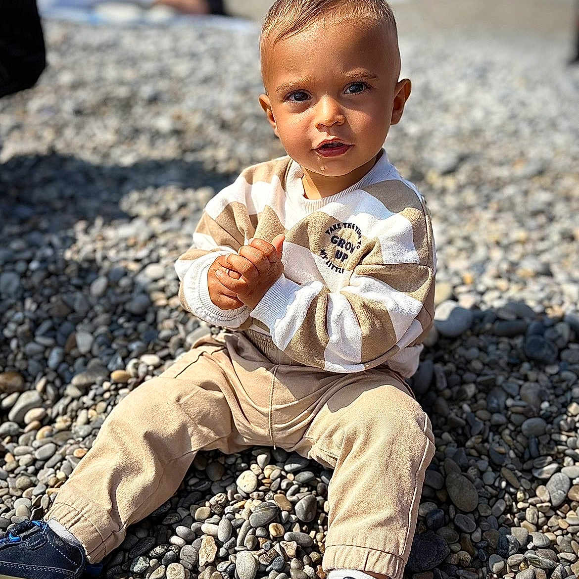Kevan a rejoint le concours — aidez-le/la à gagner de superbes lots ! background_blur, beige, casual_clothing, child, curious, cute, daylight, hands_clasped, natural_light, outdoor, pebbles, person, portrait, shoes, short_hair, sitting, sneakers, striped_sweater, toddler, young_child