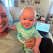 Grayson joined the competition — help win amazing prizes! baby, adult, smile, couch, living_room, laptop, onesie, colorful_clothing, hand, face, window, curtain, lamp, decor, sign, plant, table, person, indoor, furniture