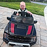 baby, child, toy_car, black_car, ford, raptor, outdoor, driveway, grass, greenery, smile, steering_wheel, pavement, suburb, house, daylight, person, vehicle, happy, front_view