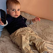 Rafaël participe au concours pour gagner de l'argent avec cette photo : toddler, child, remote_control, beige_pants, socks, bed, blanket, blue_cushion, indoor, person, casual_clothing, toy, young_child, facial_expression, lying_down, home, cozy, light, skin, hair