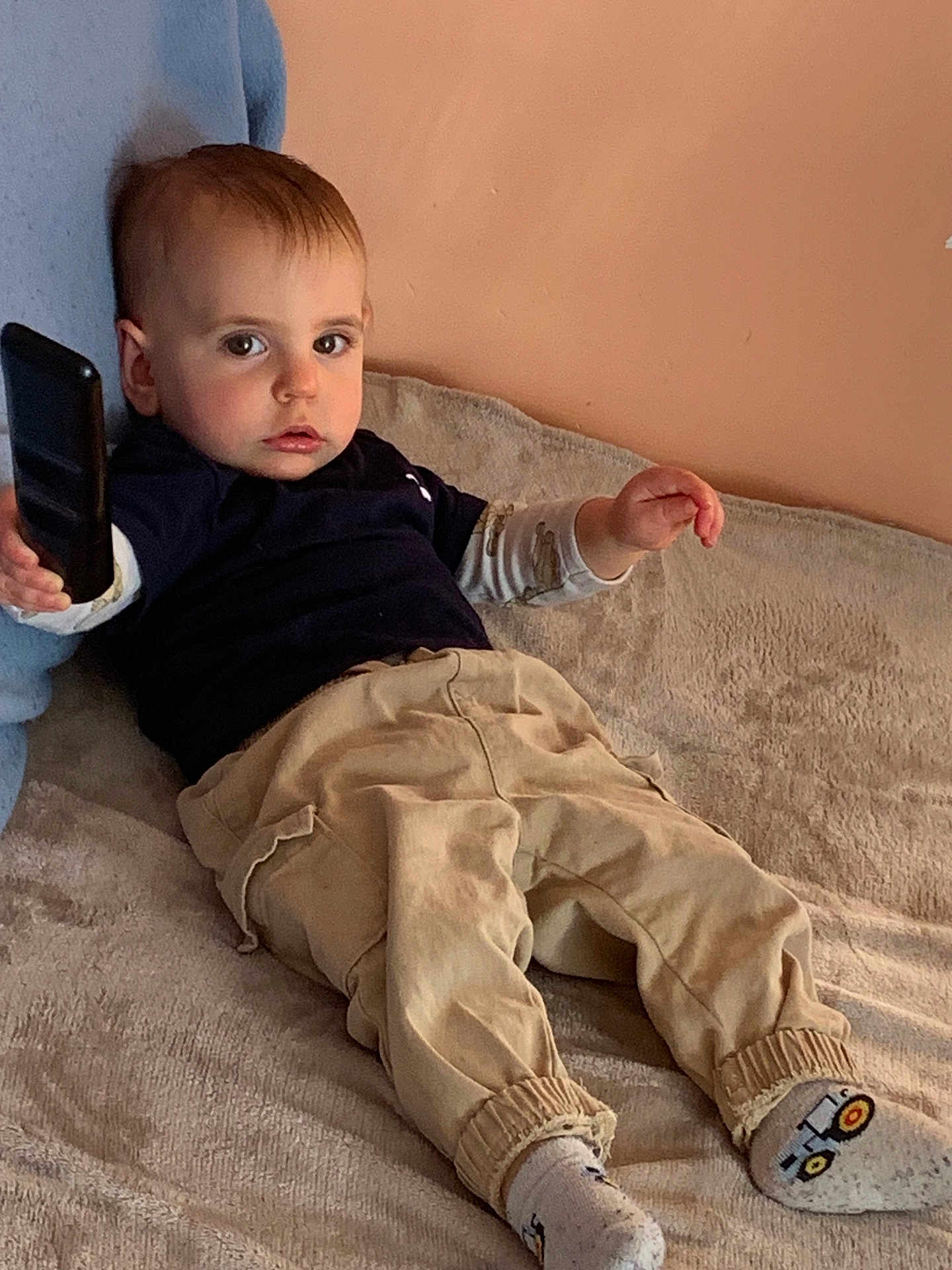 Rafaël participe au concours pour gagner de l'argent avec cette photo : toddler, child, remote_control, beige_pants, socks, bed, blanket, blue_cushion, indoor, person, casual_clothing, toy, young_child, facial_expression, lying_down, home, cozy, light, skin, hair