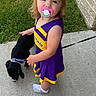 toddler, child, pacifier, puppy, dog, leash, sidewalk, grass, brick_wall, purple_dress, yellow_trim, outdoor, curious, cute, small_dog, person, footwear, sock, pet, summer