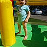 Gideon is registered to the contest to win money with this photo: toddler, curly_hair, barefoot, inflatable, bounce_house, yellow_pillar, blue_shirt, outdoor, sunlight, shadow, grass, trees, cars, mesh, play, child, smile, fun, summer, green_floor