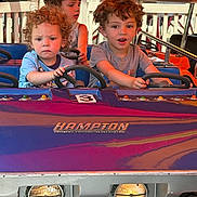 Gideon is registered to the contest to win money with this photo: child, children, amusement_ride, car, steering_wheel, curly_hair, short_sleeves, outdoor, daylight, play, fun, kids, face, vehicle, seat, smile, concentration, colorful, hands, young