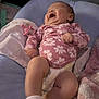 baby, infant, smiling, laughing, onesie, pink_clothing, floral_pattern, diaper, white_socks, cushion, blanket, plush_toy, penguin, indoor, soft_lighting, comfort, happy, cute, child, portrait