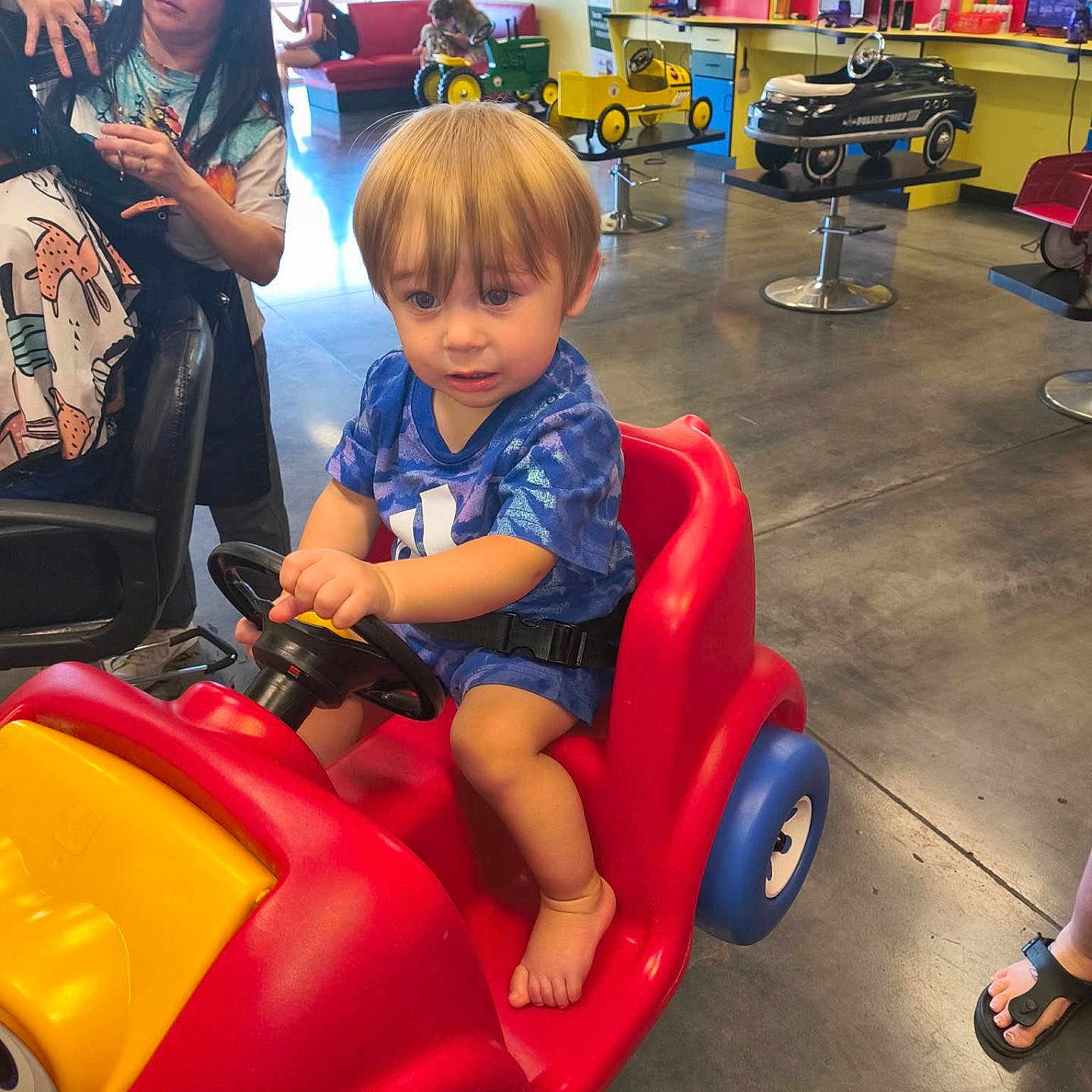 Thomas is registered to the contest to win money with this photo: toddler, toy_car, child, haircut, salon, woman, indoor, floor, colorful, reflection, pedestal, playful, curious, steering_wheel, blue_clothing, red, yellow, black_hair, footwear, person