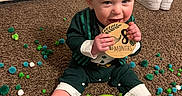 Thomas is registered to the contest to win money with this photo: baby, child, carpet, pom_poms, toy, plaque, sitting, smiling, indoor, clothing, barefoot, green, white_shoes, floor, door, happy, cute, infant, playful, portrait
